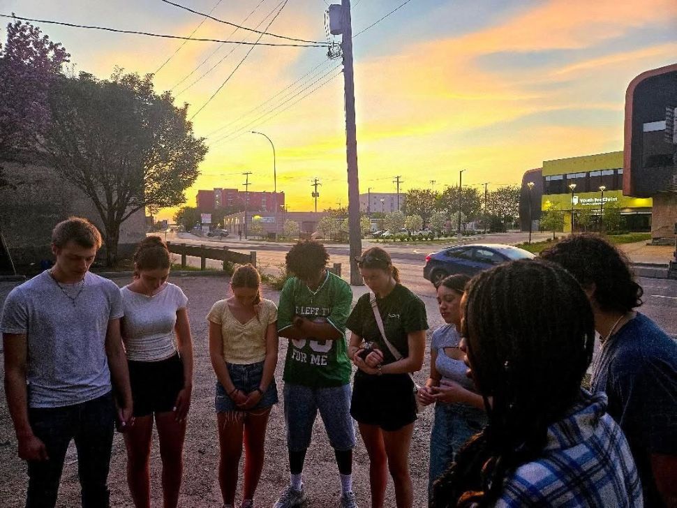 young adults bowing heads in prayer with sunset in background